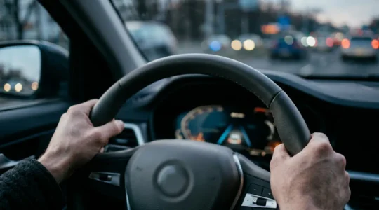 Driver's hands gripping steering wheel inside modern vehicle with subtle dashboard lights indicating active driver assistance systems