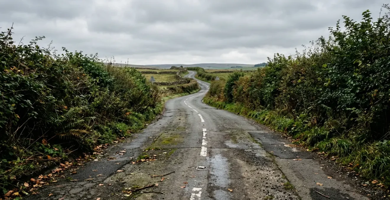 Narrow British B-road with faded white lane markings, hedgerows casting shadows, and tight curves illustrating challenging conditions for lane keeping systems