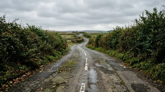 Narrow British B-road with faded white lane markings, hedgerows casting shadows, and tight curves illustrating challenging conditions for lane keeping systems