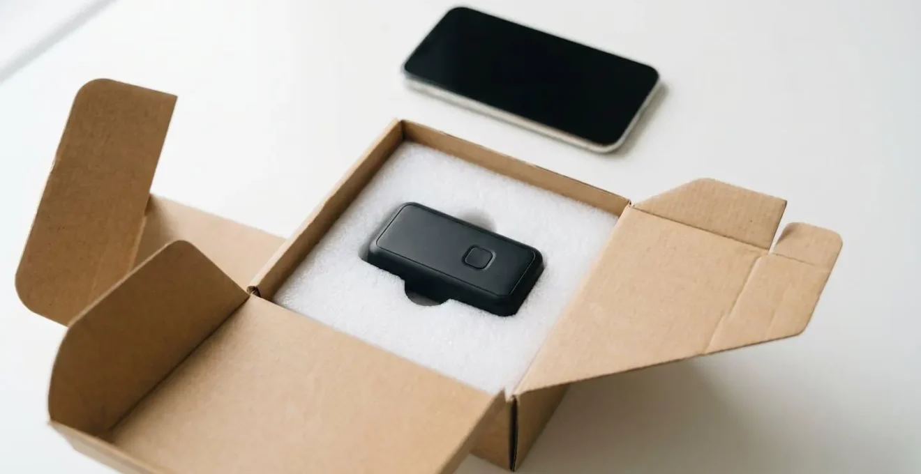Overhead view of compact GPS tracking device placed inside protective packaging foam with smartphone showing location map in background